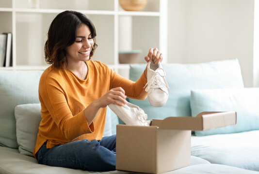 Portrait of happy young arab woman unboxing parcel with shoes at home, smiling middle eastern female opening cardboard box and looking at new pair of white sneakers, satisfied with online shopping