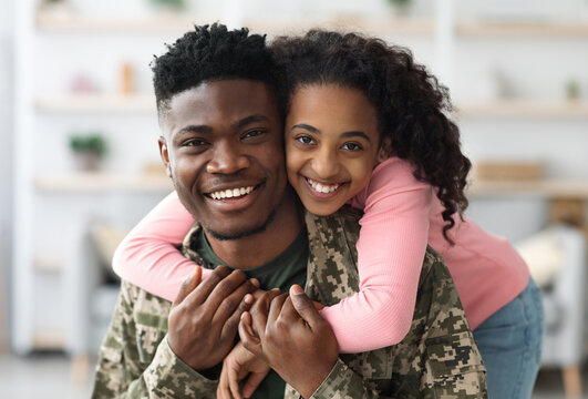 Fototapeta Portrait of cheerful pretty curly black teen girl hugging her dad soldier from behind and smiling at camera, young black man in military uniform return home from army, cuddling with his daughter