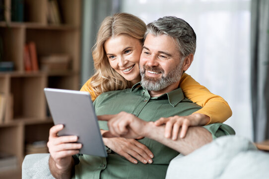 Happy Middle Aged Couple Using Digital Tablet While Relaxing In Living Room Together, Smiling Mature Man And Woman Shopping Online, Browsing Internet Or Checking New Application, Closeup Shot - Powered by Adobe