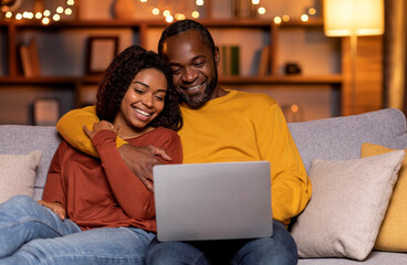 Happy smiling african american spouses sitting on couch in living room decorated with lights, bonding, using computer laptop, black man and woman shopping online from home, copy space