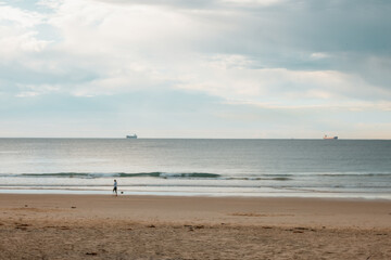 Person with motion blur walking along beach with cargo ships on the horizon
