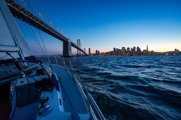 A sailboat cruises on the water at dusk, passing under the Bay Bridge with the San Francisco...