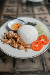A plate of Thai chicken rice with fresh tomatoes, cucumbers, and spicy dipping sauce, served on a rustic wooden table.