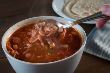 Spoon taking charro beans from a white bowl with a plate with flour tortillas next to it and with a gray napkin on the table. Mexican food