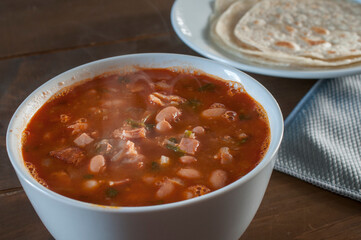 Charro beans in a white bowl on a wooden table with flour tortillas and a gray napkin next to them.