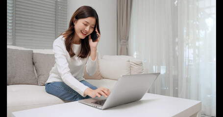 Asian woman is talking on the mobile phone and using laptop computer in the living room at home