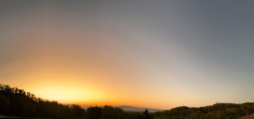 Wide panoramic sunrise over hills with glowing orange sky and scattered trees, creating a dramatic and peaceful landscape view.