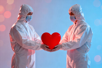 Two medical professionals in protective suits holding a red heart together against a soft blue background.
