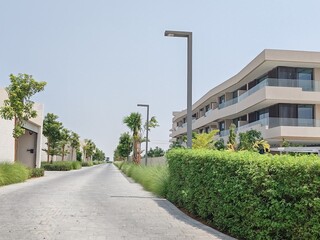 Fototapeta premium Interlocking pathway with ramp road runs through landscaped area, featuring greenery trees on both sides. Houses or villas are situated beside pedestrian ramp and stairway, all set under a cloudy sky.
