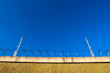A concrete wall topped with razor wire and an electric fence stands against a clear blue sky,...