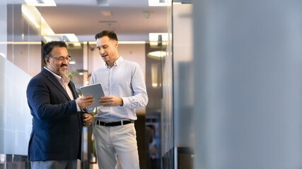 Two professionals stand in office hallway, discussing project on tablet