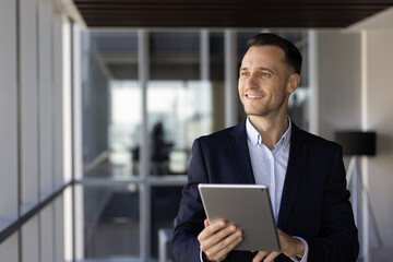 Fototapeta premium Optimistic Hispanic businessman holding digital tablet, stands in office hallway