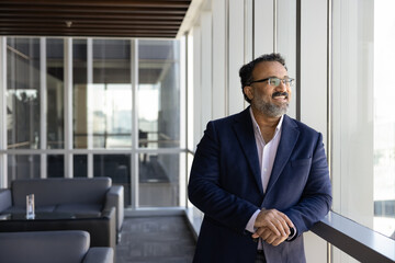 Fototapeta premium Middle-aged businessman stands by office window looking outside with smile
