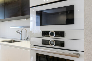 Fototapeta premium A modern, minimalist kitchen featuring built-in white ovens and microwave. Stainless steel sink and faucet visible on the left. Clean lines and bright lighting