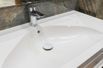 A close-up of a clean, white bathroom sink with a chrome faucet. The background features stone-like tiles