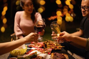 Concept for friendship, celebration, and lifestyle. A group of friends making a toast with various drinks wine, beer, and spirits during an outdoor night gathering.