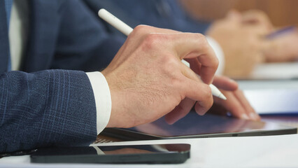 Male lawyer’s hands in a suit use a stylus on a digital tablet during an office meeting; smartphone nearby, modern workflow with e‑sign, notes and document review. Photo