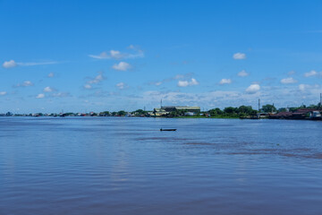 Kapuas River stretches wide under the bright blue sky, gracefully dividing Pontianak city into two sides, with houses and boats lining its calm brownish waters.