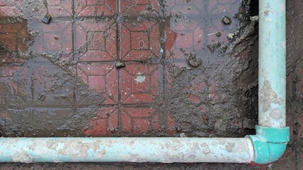 A closeup of a wet, dirty tile floor with a blue pipe forming a corner, showcasing the effects of water damage and neglect in an outdoor setting