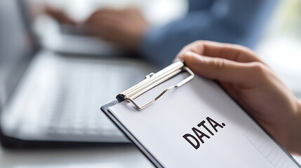 Analyzing Data: Person holding a clipboard with the word "DATA" in front of a laptop, symbolizing data analysis and collection in a modern workspace.