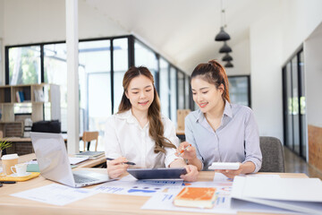Two businesswomen discussing work using digital tablet and laptop in modern office, Business team analyzing financial data on a digital tablet and paper report.