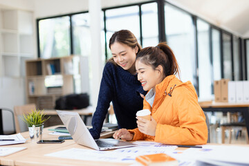 Fototapeta premium Two cheerful diverse office workers women cooperating on project, sitting at work desk with laptop, looking at screen, smiling.