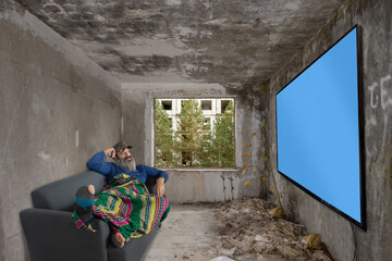 Bearded man lying on couch in abandoned room and watch TV on wall with blue screen