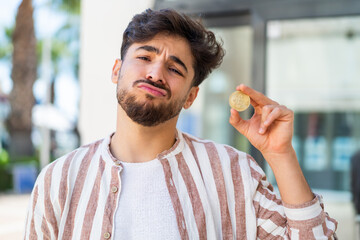 Handsome Arab man holding a Bitcoin at outdoors with sad expression