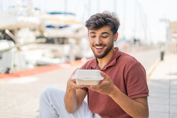 Young Arabian handsome man at outdoors taking a box of takeaway food with happy expression