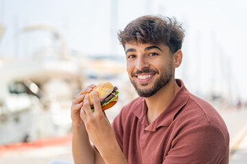 Young Arabian handsome man at outdoors holding a burger with happy expression
