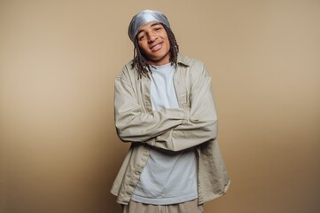 Young person with braided hair wearing a headscarf and casual clothing, standing with arms crossed against a neutral background.