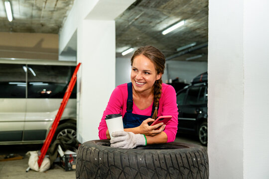 Female auto mechanic taking coffee break in auto repair shop, using smartphone and drinking coffee.
