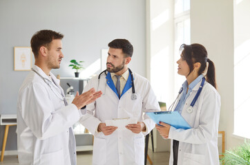 Fototapeta premium Three doctors engage in focused discussion in medical office during busy workday. Male and female medical workers in white coats talk, exchanging ideas about patient care. Medicine concept.