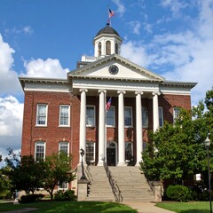 Obraz premium A majestic brick building with a classical design, featuring a prominent portico and a cupola, stands on a landscaped lawn under a bright blue sky