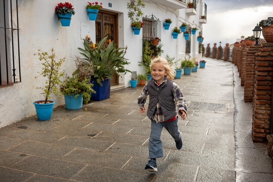 Happy little blonde boy running along a charming narrow street with flowerpots on white walls and red brick fence after rain in a picturesque Andalusian village during golden hour.