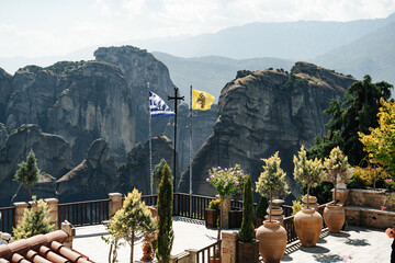 Byzantine flags waving in the wind, dramatic rock formations of Meteora rise steeply, creating a striking contrast between nature and human architecture. large terracotta pots, trees, and Greek 