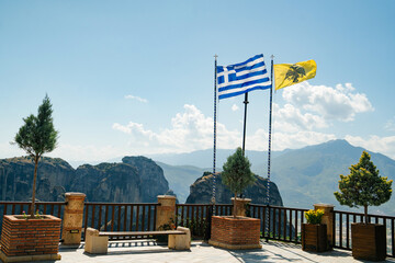 Byzantine flags waving in the wind, dramatic rock formations of Meteora rise steeply, creating a striking contrast between nature and human architecture. large terracotta pots, trees, and Greek 