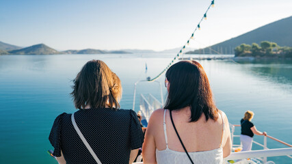 Two women standing side by side on the deck of a boat, enjoying a peaceful sea cruise on a calm, sunny day. They are looking out at the turquoise water, distant hills and marina in the background, 