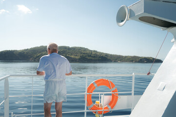 Elderly man standing on the deck of a ferry boat in Greece, looking at the calm blue Aegean Sea and the green coastline in the distance.