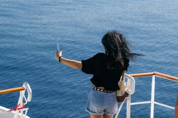 Woman with long dark hair, wearing a black blouse and denim shorts, takes a selfie on the deck of a ferry boat. The deep blue sea stretches endlessly in the background, evoking a sense of freedom