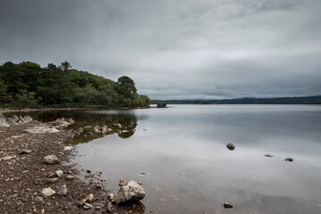 A tranquil scene at Lough Leane, Ireland, with a rocky shoreline, calm water reflecting the cloudy sky, and a dense treeline along the lake’s edge