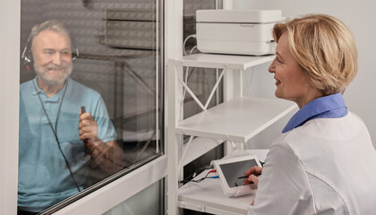 Hearing testing. Senior male patient wearing audiology headphones pressing button of response while audiometric testing in soundproof audiology booth