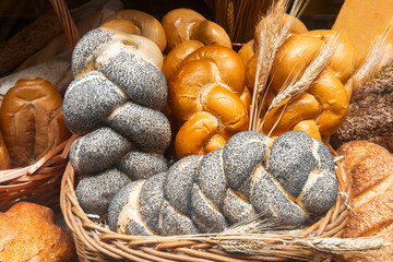 Various types of freshly bread in a wicker basket and wheat ears, closeup. Braid buns with poppy seeds for baking. Pastry background.