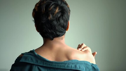 Person With Skin Condition Suffering From Itch Scratching Their Back in Blue Off Shoulder Top Against Plain Backdrop Demonstrating Discomfort and Seeking Relief With Dark Hair Style