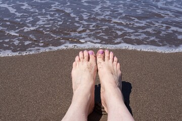Woman’s feet with pink toenails in sand near ocean. Relaxation and beach lifestyle concept with close-up view of feet on wet shoreline as sea waves approach. Perfect image for summer vacation themes.