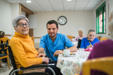 Obraz premium Caregiver assisting senior woman in wheelchair at nursing home, other elderly residents in the background enjoying activities