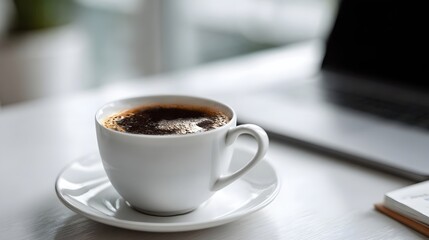 White cup of black coffee on a desk next to a blurred laptop