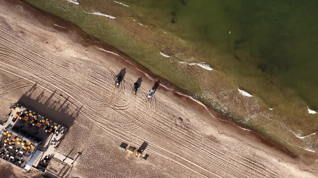 Top-down aerial view of a sandy beach with a beachfront caf&eacute;, outdoor seating, and waves gently washing ashore in Palanga, Lithuania.
