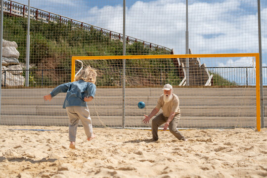 Barefoot child kicking soccer ball towards elderly man acting as goalkeeper on sandy beach field, concept of intergenerational activity, family bonding, summer sport and outdoor fun.