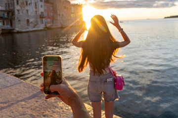Girl posing at sunset while being photographed by phone. A teenage girl poses playfully in golden sunset light by the sea, while someone captures the moment on a smartphone.